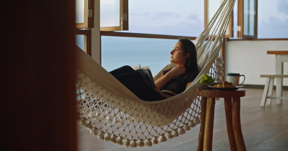 A woman is relaxing on a hammock holding a book and looking out the window as she gently rocks back and forth