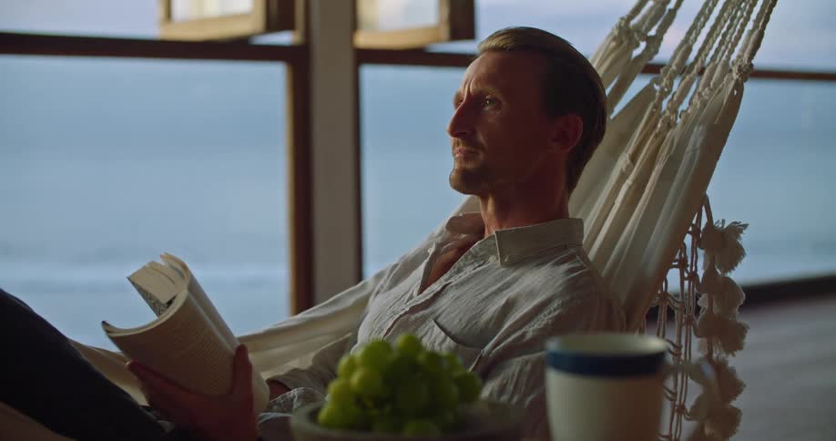 A man is relaxing on a hammock reading a book then he closes the book and leans his head back and closes his eyes