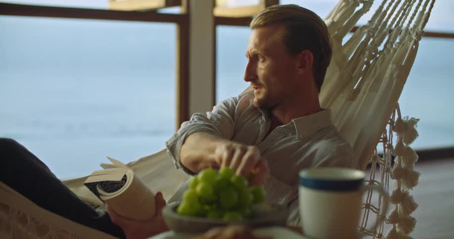 A man is relaxing on a hammock reading a book and he reaches over to the table and takes a green grape and puts it in his mouth