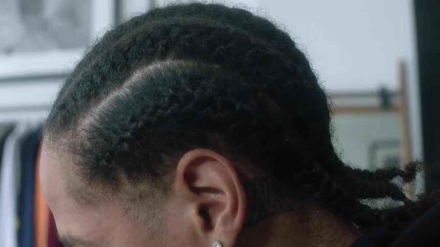 A man has cornrows in his hair and there is a rack of clothes in the distance