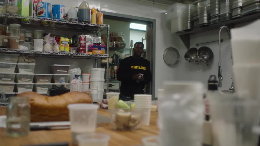 A man is standing in the doorway of a restaurant kitchen looking at bread and containers on a table as he wipes his hands with a towel