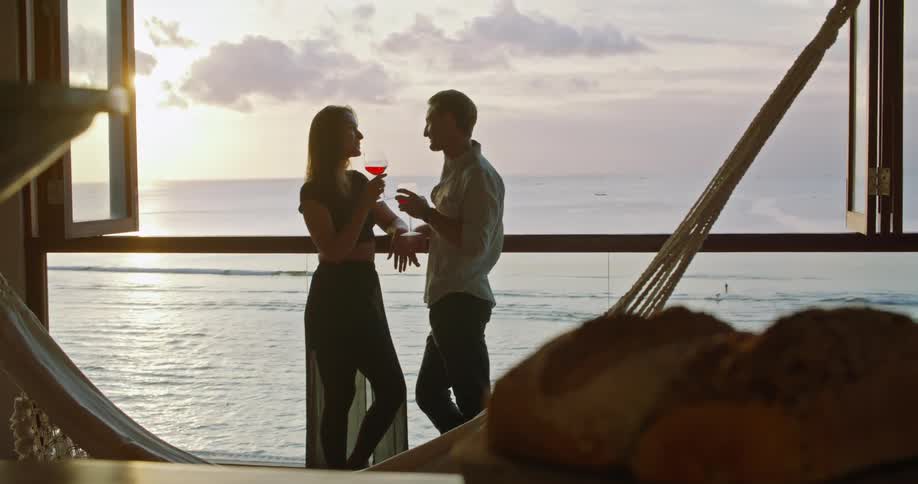 A man and woman kiss and drink wine while leaning against a large window overlooking the sea in the evening