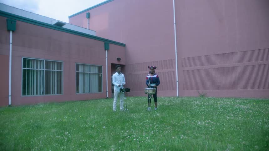 A boy and a girl are standing in the grass outside of the school building playing snare drums