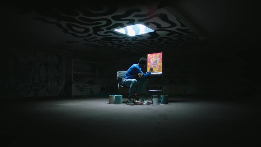 A man sitting in the center of a room directly under a skylight painting a canvas