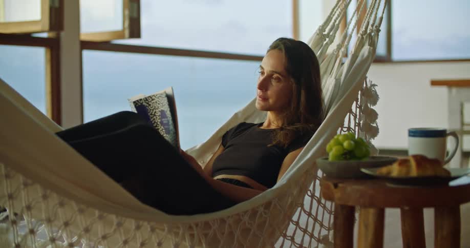 Woman reads in swaying hammock placed between stool topped with food and open windows