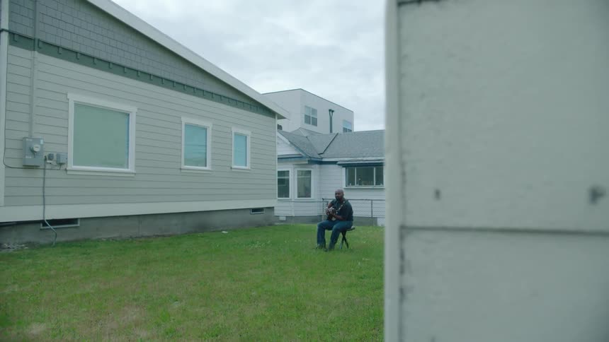 Man sitting on a chair in the grass in front of a house playing acoustic guitar