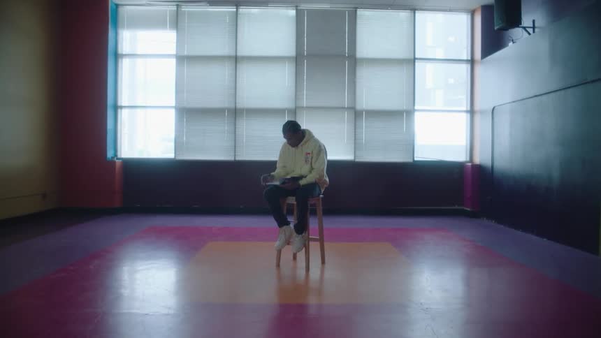 Man sitting on a tall stool in the middle of an empty room with colorful flooring writes on a notebook