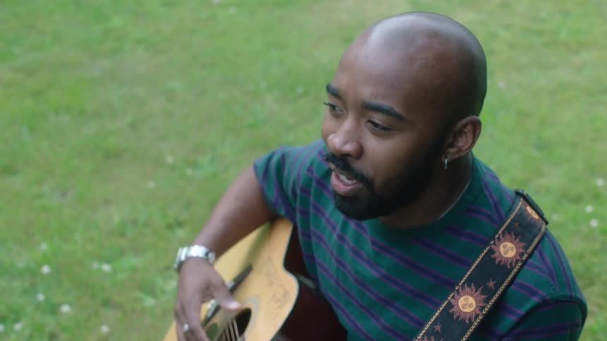 Bald man with earrings beards blue striped shirt and silver wriswatch plays guitar sitting on green grass in public park