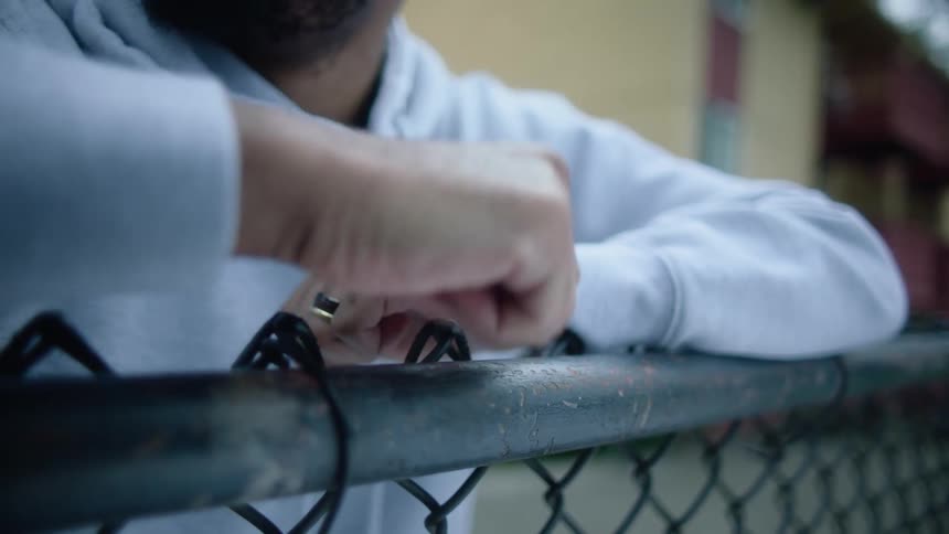 Man reclined on a wired fence tapping the top with his knuckles near a building