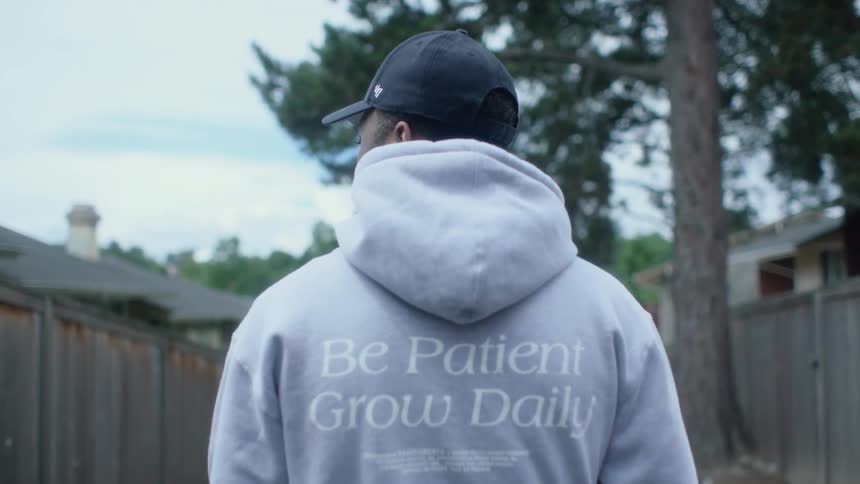 Man with blue cap and gray hoodie stands in garden looking sideways on cloudy day