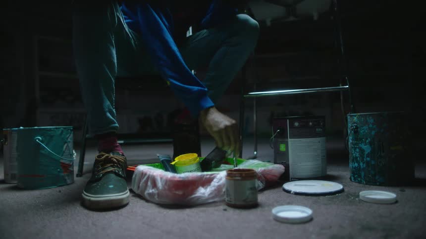 Someone is sitting on a chair and paint cans are on the floor at their feet as they bend down and dip their paintbrush into the cans