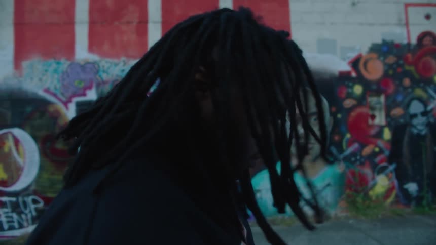 Man with dreadlocks and black jacket shakes his head next to wall with graffiti