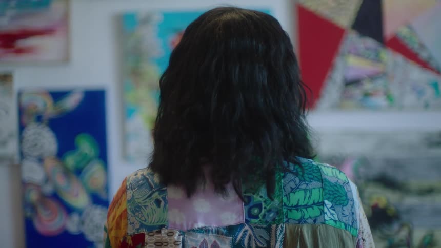 A woman with black hair has her back turned and she is facing a wall with artistic pictures