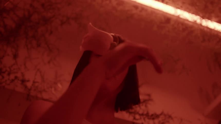 A woman holds a rose and caresses her face with it while in a room vines and a reddish glow