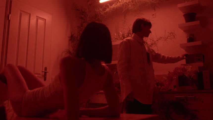 In room lit by red colored lighting man stands by shelves and turns on machine as woman reclines