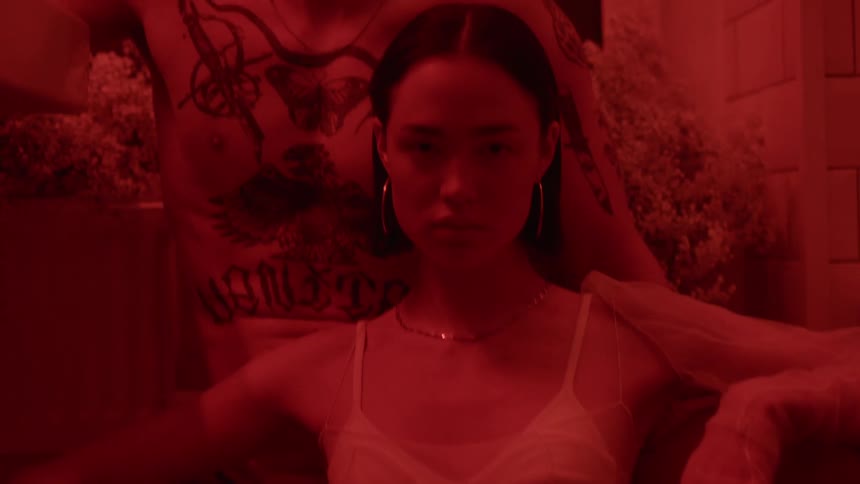A man and woman gaze into the camera while posing in sitting position in their underwear in a room illuminated by red lighting