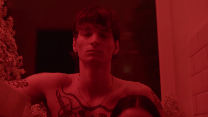 A man is standing in a room illuminated with red light and he has tattoos on his chest