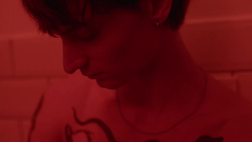 A young person leaning against a tile wall head nodding forward bathed in a reddish glow