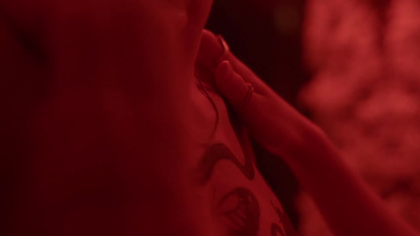 Woman touches neck and shoulder of tattooed man in room lit by red light
