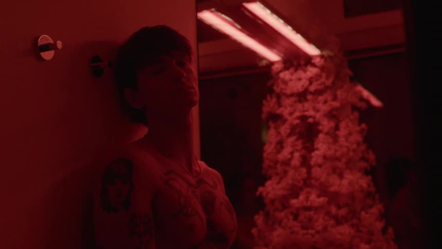 A shirtless man looks into the camera while leaning against a wall in a room illuminated by red lighting
