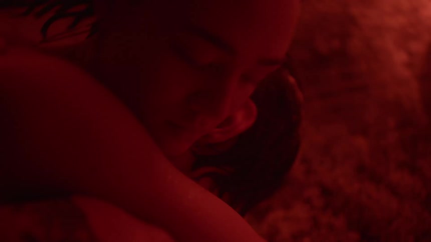 A man and a woman are lying down together in a room illuminated with red light and her arms are around his neck