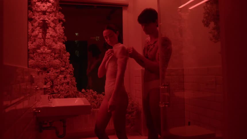 Man helps woman adjust sleeve as they stand in bathroom at night under red colored lighting