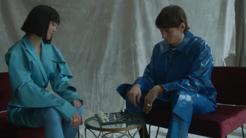 A man and woman in fashionable clothing play chess while sitting at a small table in a studio while being photographed