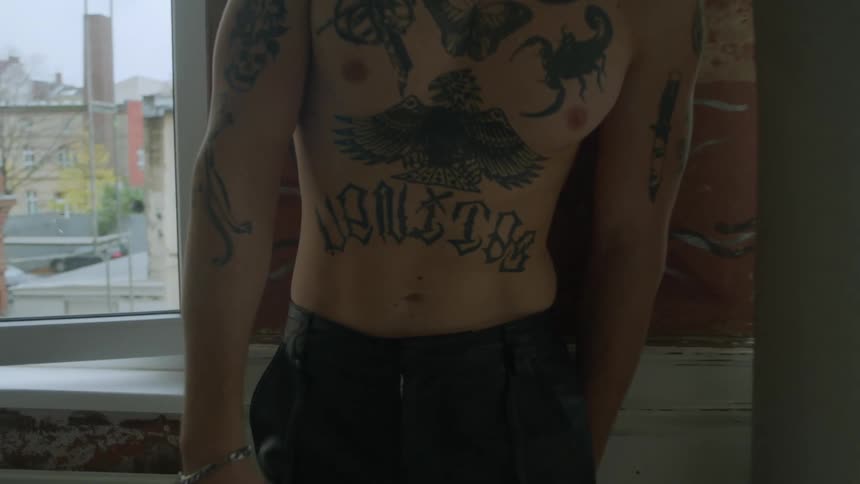 Man with tattooed chest stands near wall between two windows showing neighborhood