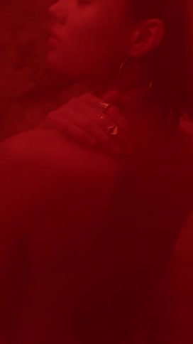 A red light shines on a womans bare back and face as she rubs her shoulder with her hand