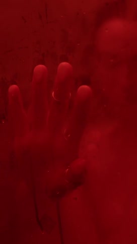 A man is in a room illuminated with red light and he is pressing his hand on a window