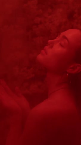 Under a red light a woman is standing in a shower and she lifts her hands to catch the falling water and then places them back on her shoulders