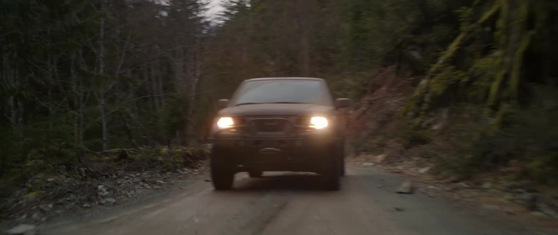 An automobile with headlights on travels on a dirt road through a forest