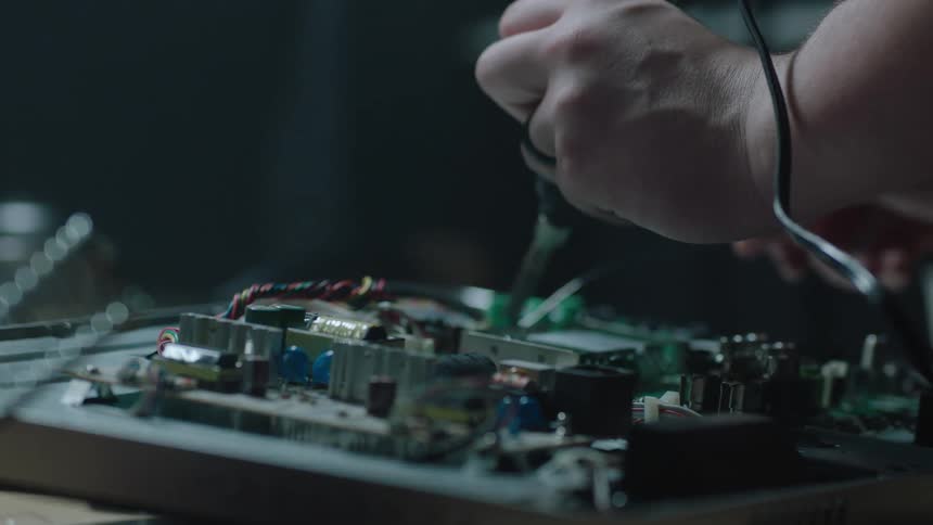 A hand reaches for a soldering iron and uses it to work on a circuit board