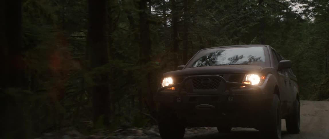 A truck travels on a mountain road through the trees with headlights on