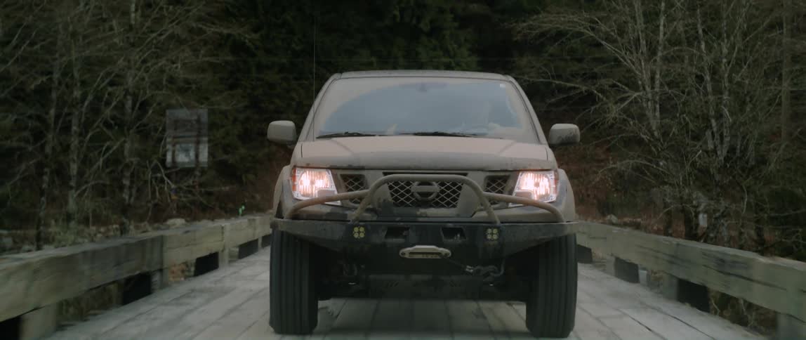 A black truck with the headlights on is traveling across a wooden bridge coming out of the woods