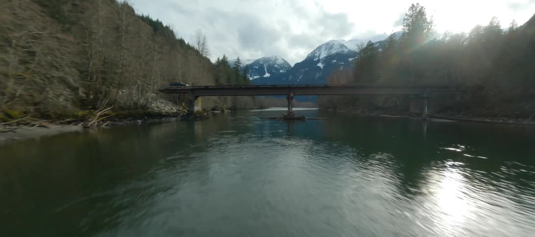Water sparkles as a truck travels over a bridge surrounded by trees and there are snow covered mountains in the distance