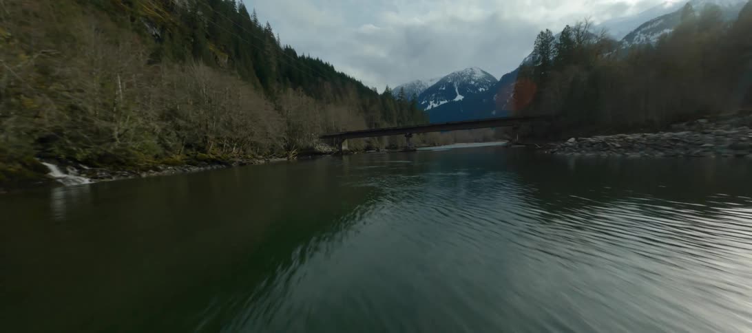 A truck drives across a bridge over a river next to a forest with mountains in the distance