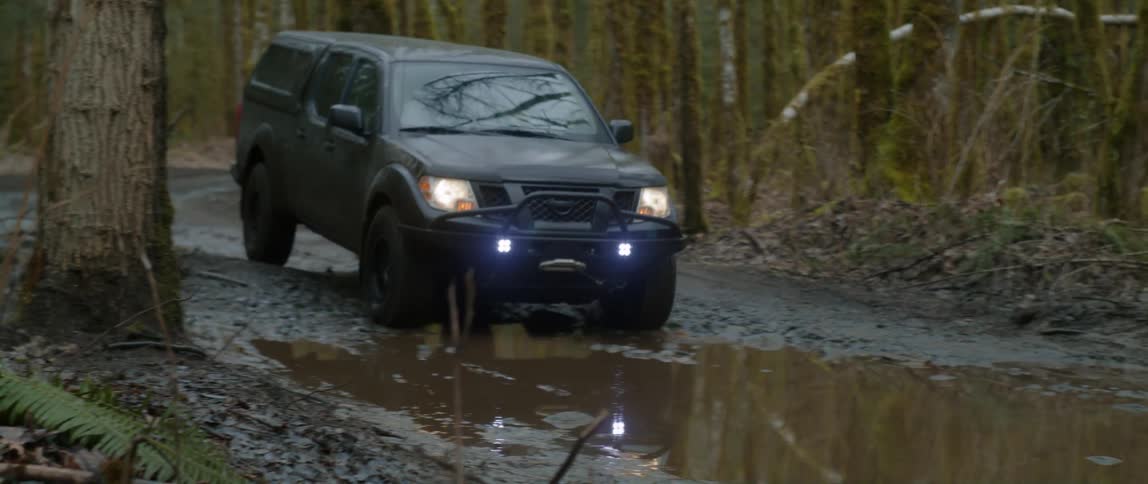 An automobile with the headlights on splashes water as it goes through a puddle on a road through the woods