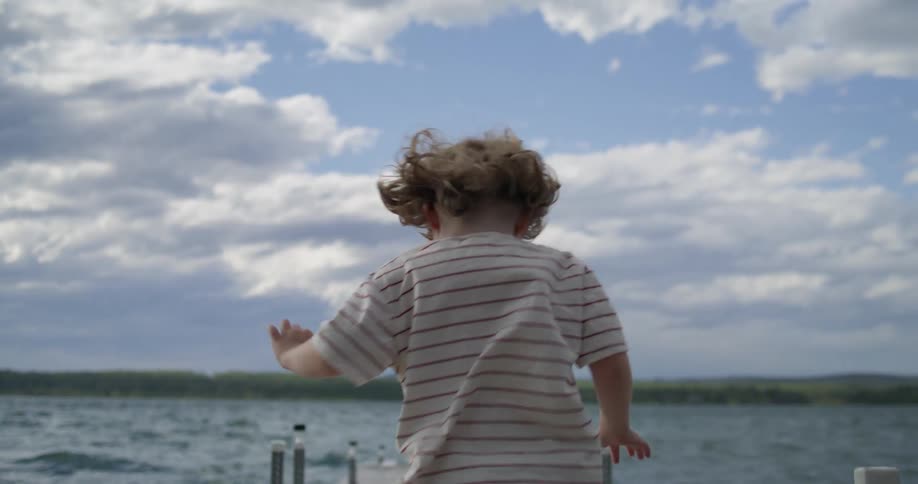 A child runs on a wooden dock with arms outstretched and hair blowing in the wind