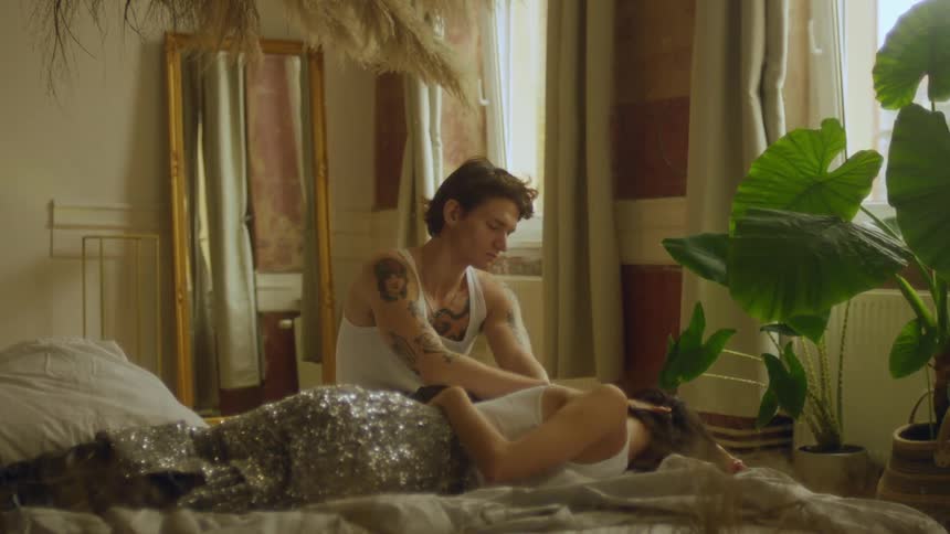 A man sits on a bed and looks down at a woman lying beside him