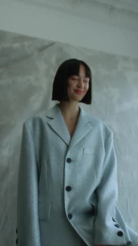 A woman smiles as she stands with her foot on a stool modeling oversized clothes and there is a sheet on the wall behind her