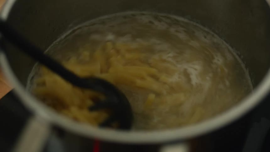 A pot of boiling water with pasta in it sits on the stove and someone puts a ladle in and stirs it