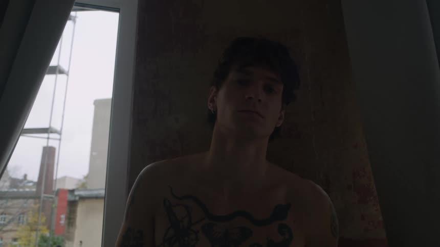 A shirtless tattooed man turns away from a window to look into the camera as a light flashes