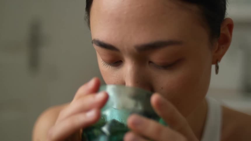 A woman is holding a glass with both hands and blows in it before she takes a drink
