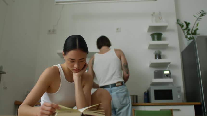 A woman is sitting at a kitchen table reading book and a man is standing at the sink