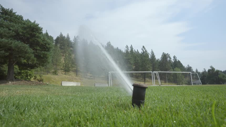 A sprinkler is spraying water onto the grass of a field near the woods as a truck passes by on the road
