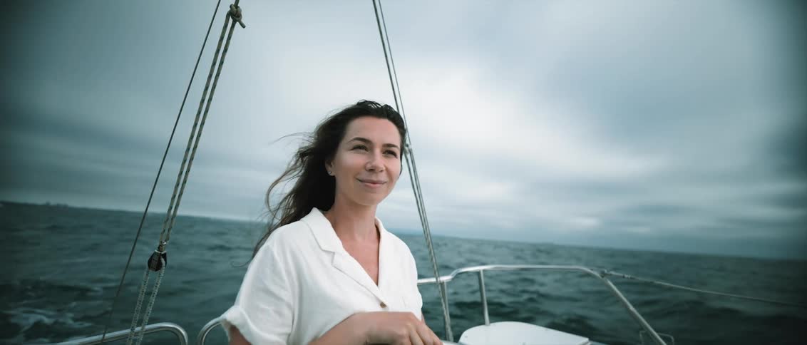 Woman with wavy hair white shirt and shorts steers a yacht on a cloudy day
