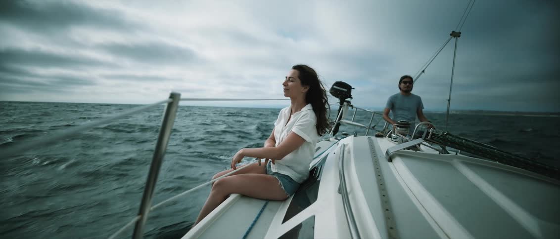 Woman with wavy hair and shorts sits on board while man with long hair and glasses drives yacht on ocean