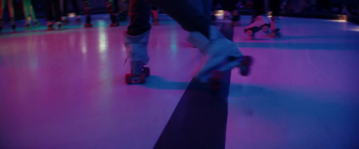 Following a young man dancing while roller skating on a rink filled with people under bright neon lights