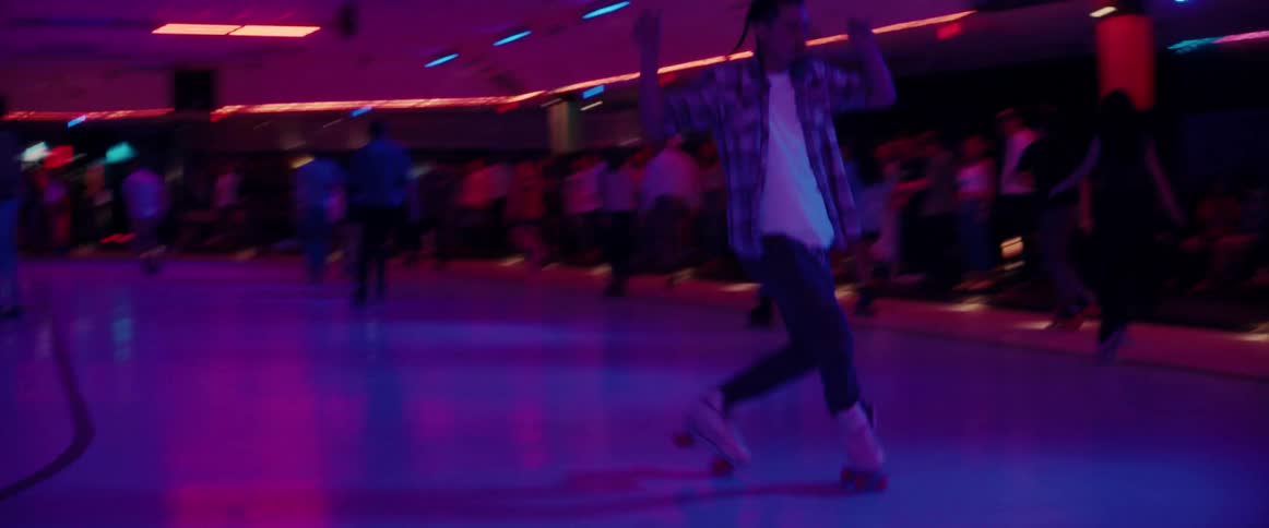Following a young man dancing while roller skating on a rink surrounded by people under bright neon lights